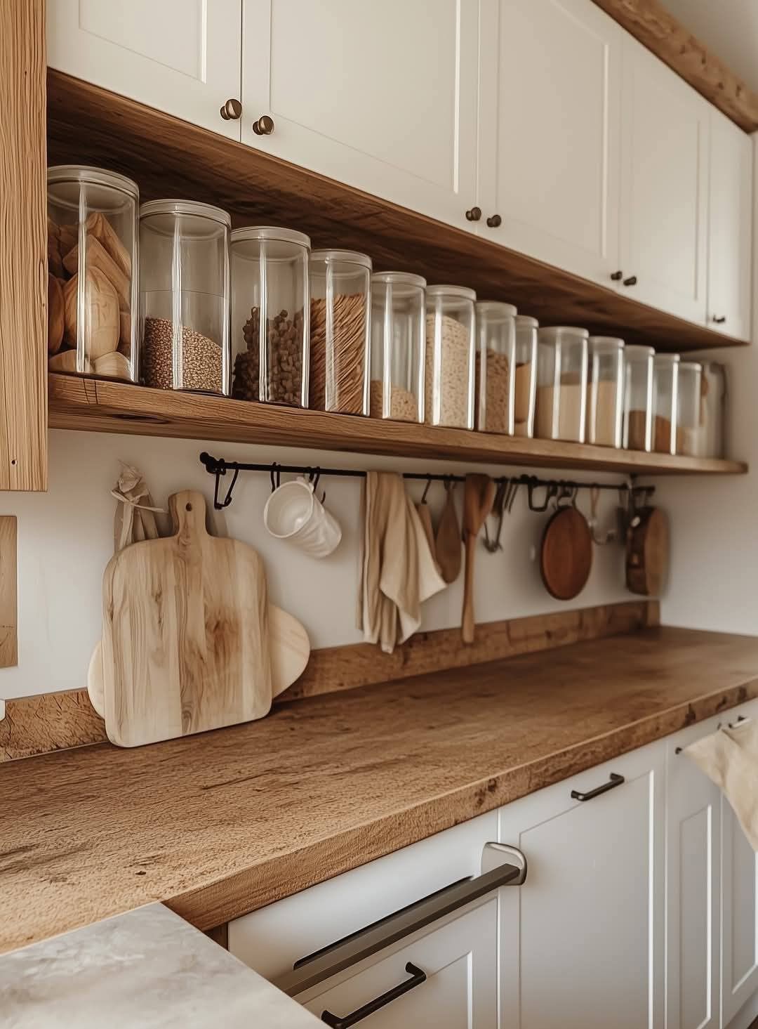 Kitchen with white upper cabinets and wooden lower cabinets. A wooden shelf holds clear canisters filled with grains and dry…