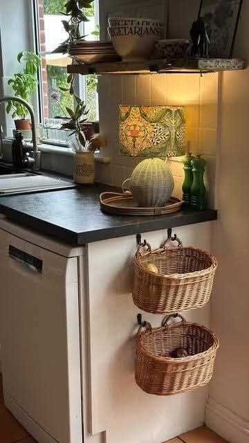 Two woven baskets hanging from hooks on the side of a white kitchen cabinet, next to a black countertop and a sink.