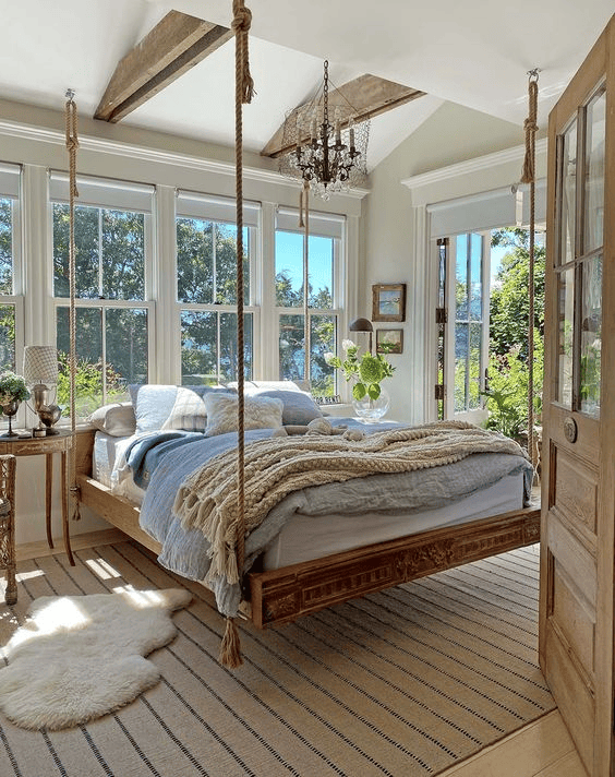 Bedroom with large windows and a natural wood hanging bed frame suspended by thick ropes, dressed in blue and white bedding.