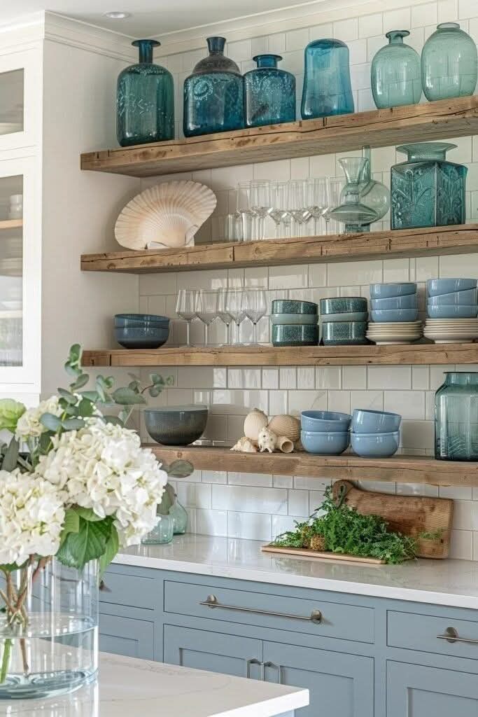 Kitchen with open wooden shelves displaying blue and green glasses, large seashell, bowls, and decorative items over subway…