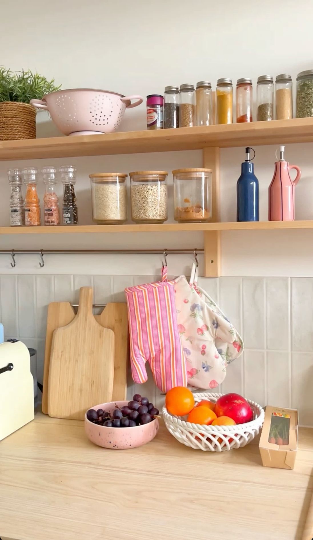 Kitchen counter with cutting boards and oven mitts hanging on hooks, open shelves with spices and grains, and a pink…