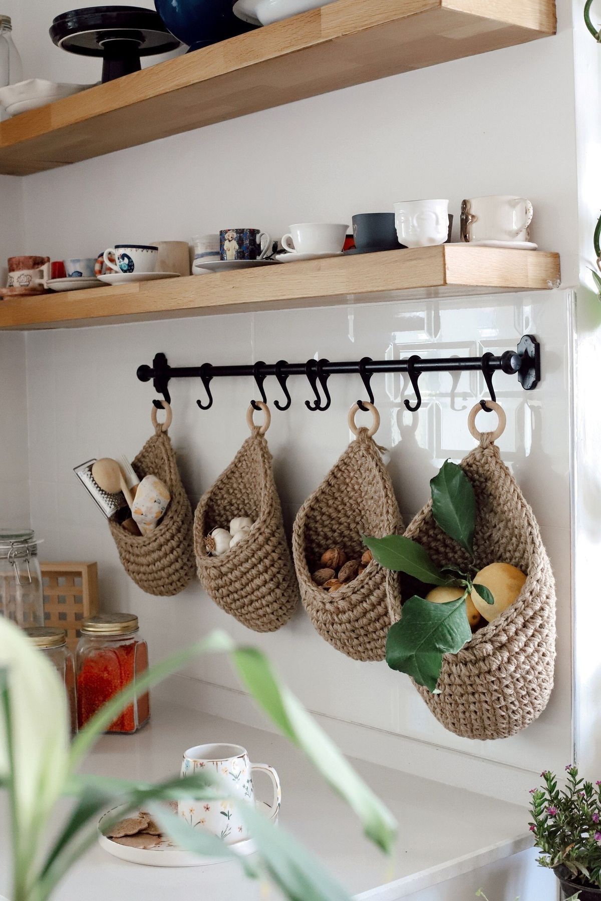 Four natural fiber hanging baskets suspended from a black rod under a shelf, holding various kitchen items and produce.