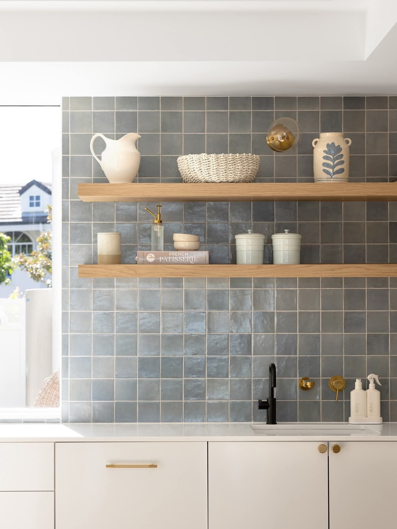 A kitchen backsplash features light blue square tiles. Two light wooden shelves hold various white and cream pottery items.