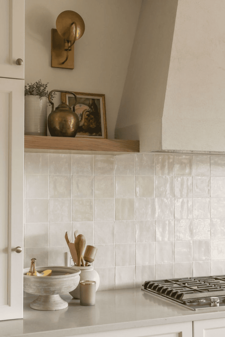 Close-up of a kitchen backsplash made of square, glossy white tiles with a range hood and open shelving above.