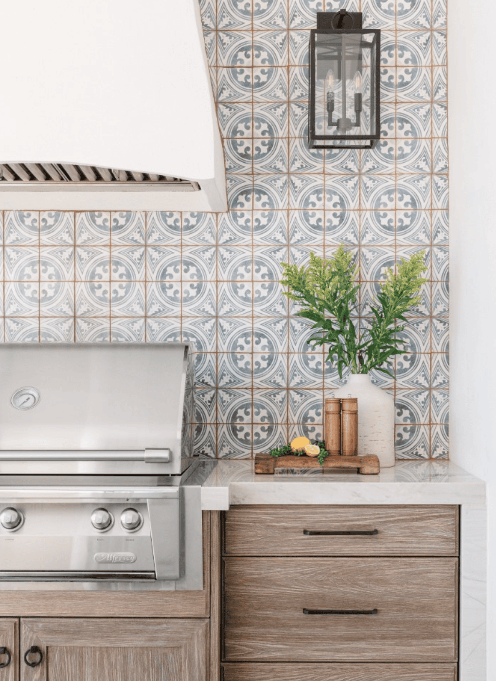 An outdoor kitchen with an ornate blue and white tile backsplash, a stainless steel grill, a white range hood, and a…