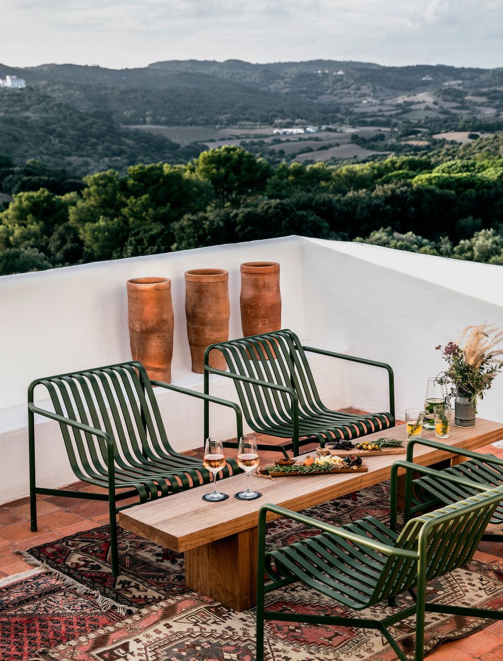 A tiled patio with four dark green slatted outdoor lounge chairs arranged around a wooden coffee table.