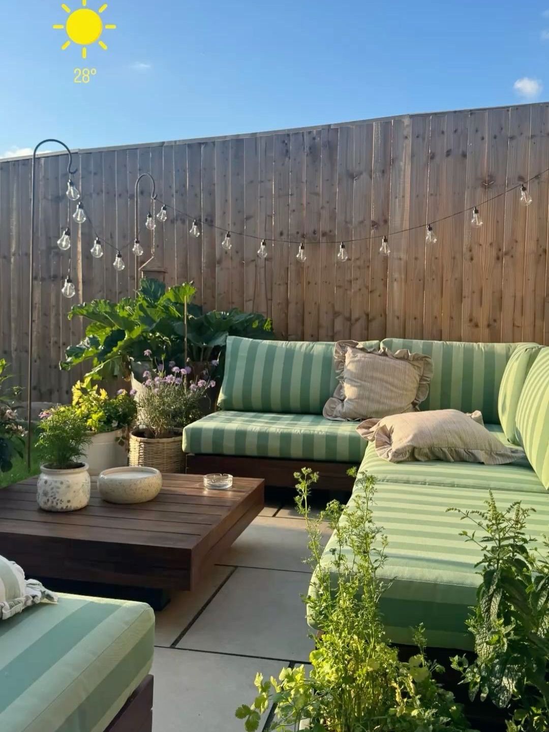 Outdoor patio with green striped sectional sofa, wooden coffee table, potted plants, and string lights hanging along a…