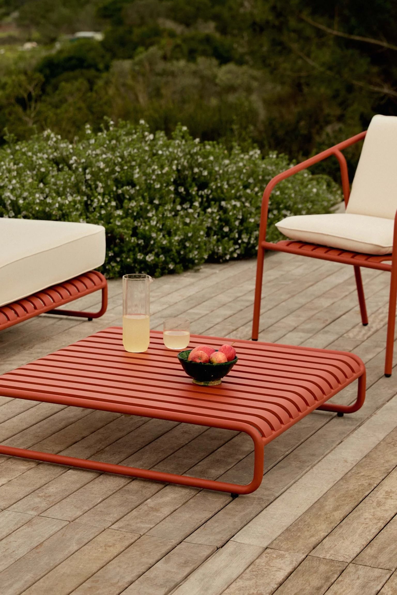 Red slatted lounge chair, red slatted coffee table with drinks and fruit, and a red dining chair with cream cushion, on a…