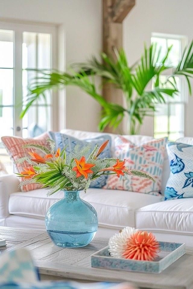 A light blue glass vase with orange daylilies on a wooden coffee table in a white living room.