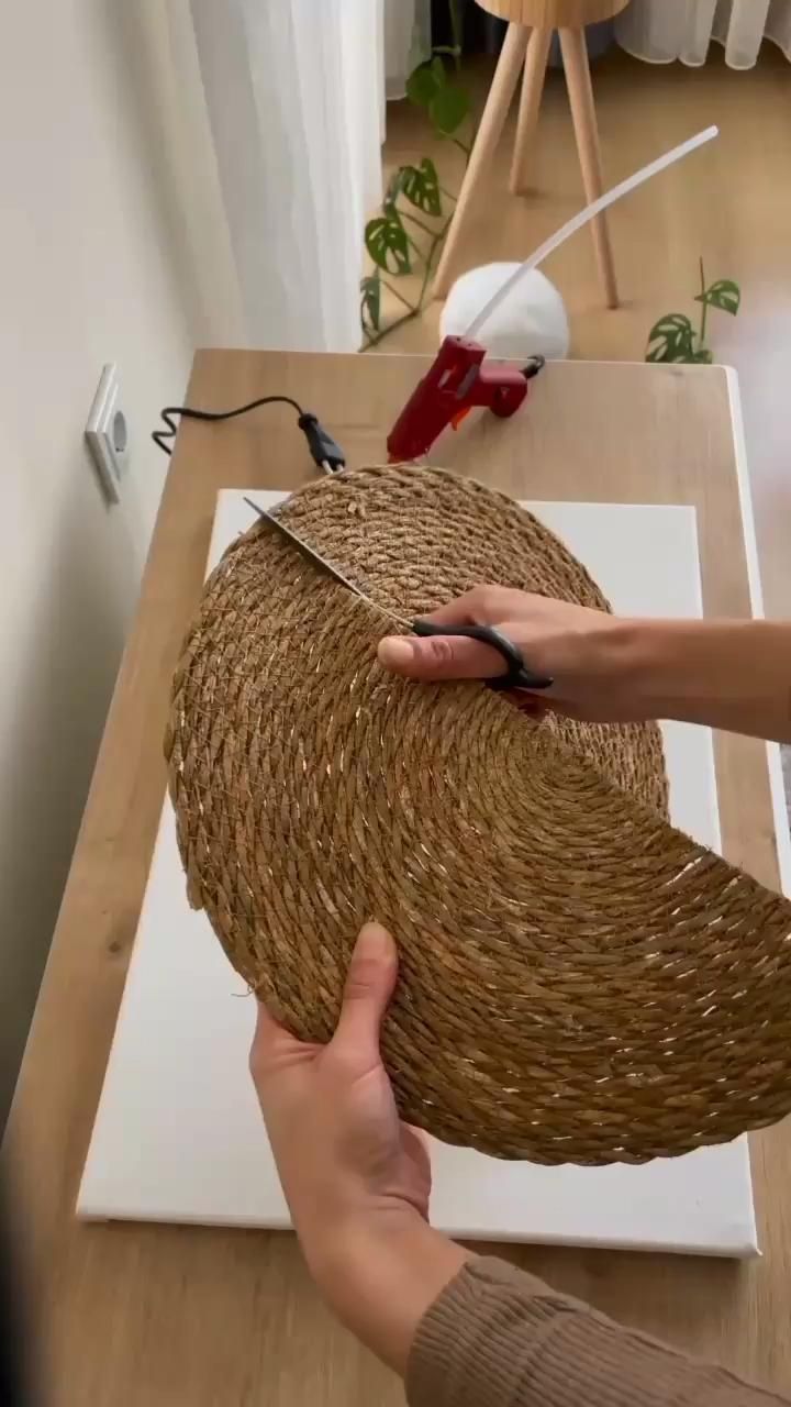 Hands cutting a round, woven fiber placemat with scissors, over a white canvas and next to a hot glue gun.