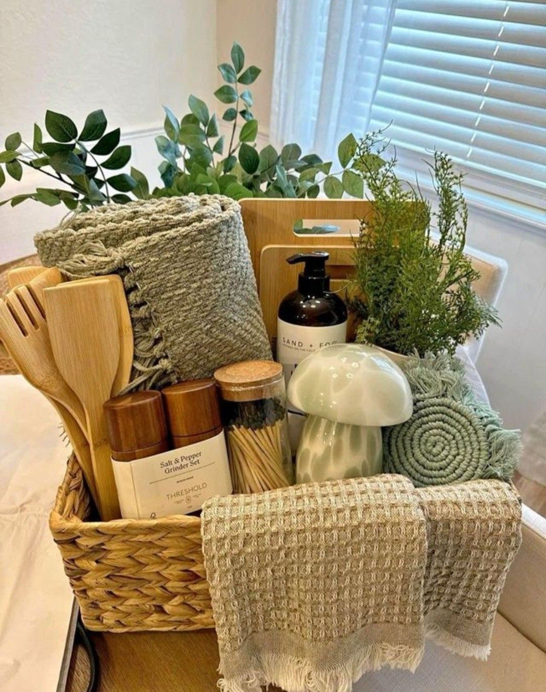 Woven basket filled with wooden utensils, salt and pepper grinders, matches, a soap dispenser, two cutting boards, and green…