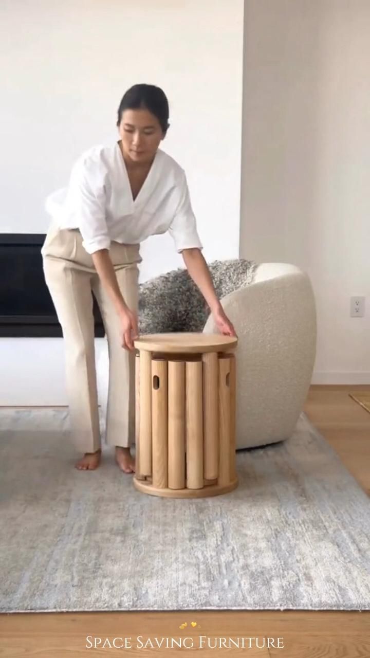 Woman in white shirt and beige pants handling a round, light wood side table that contains multiple nested stools.