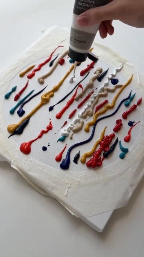 Hand squeezing paint from a tube onto a canvas with multiple colors of paint forming lines and dots.