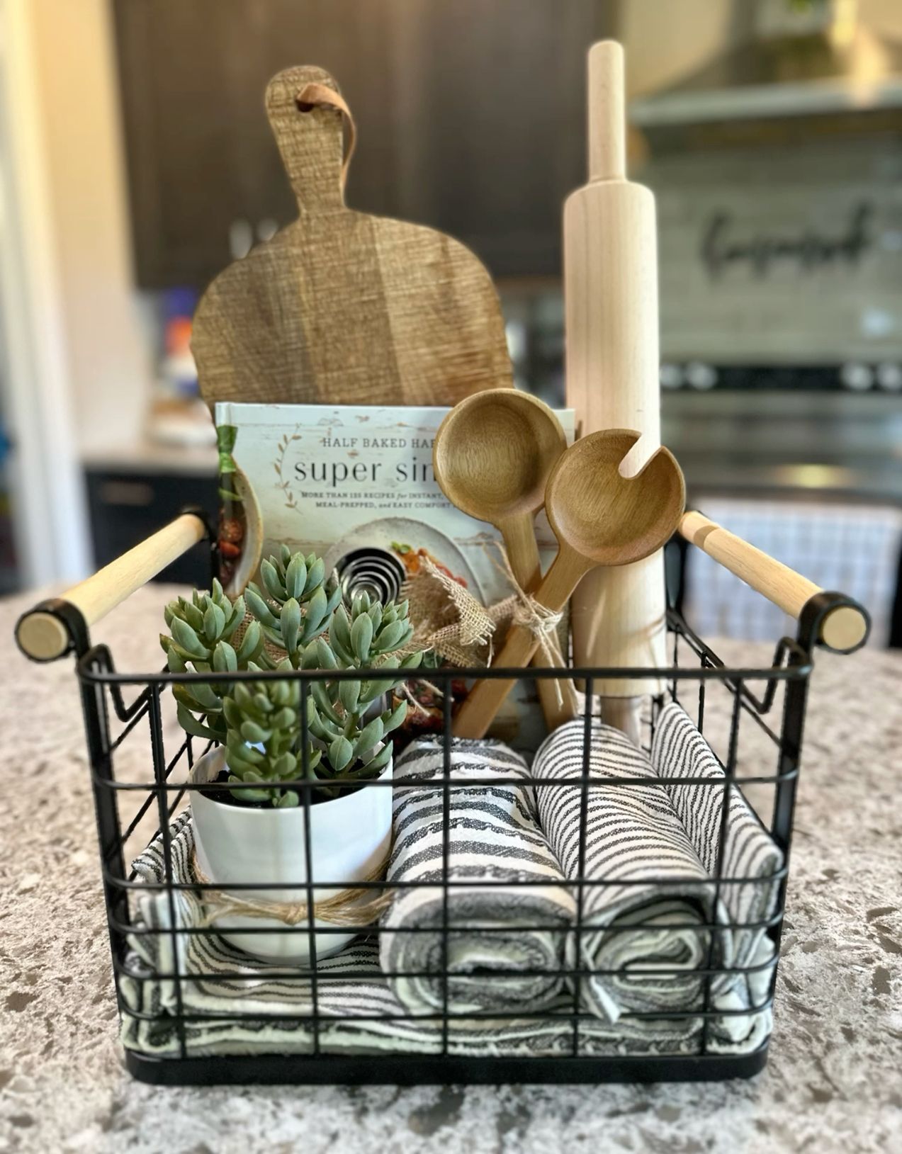 A black wire basket containing a cookbook, cutting board, wooden spoons, kitchen towels, and a small faux succulent plant.