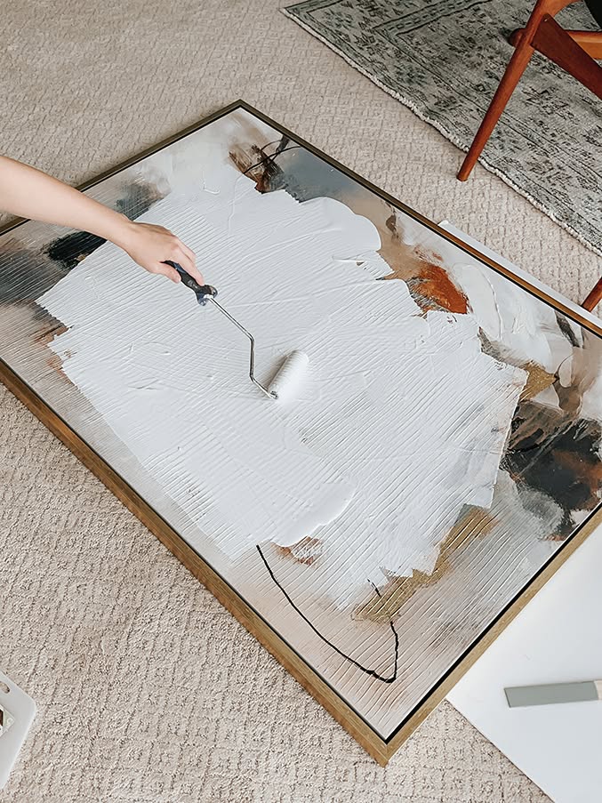 A hand rolling white plaster onto a large framed canvas, creating textured abstract art.