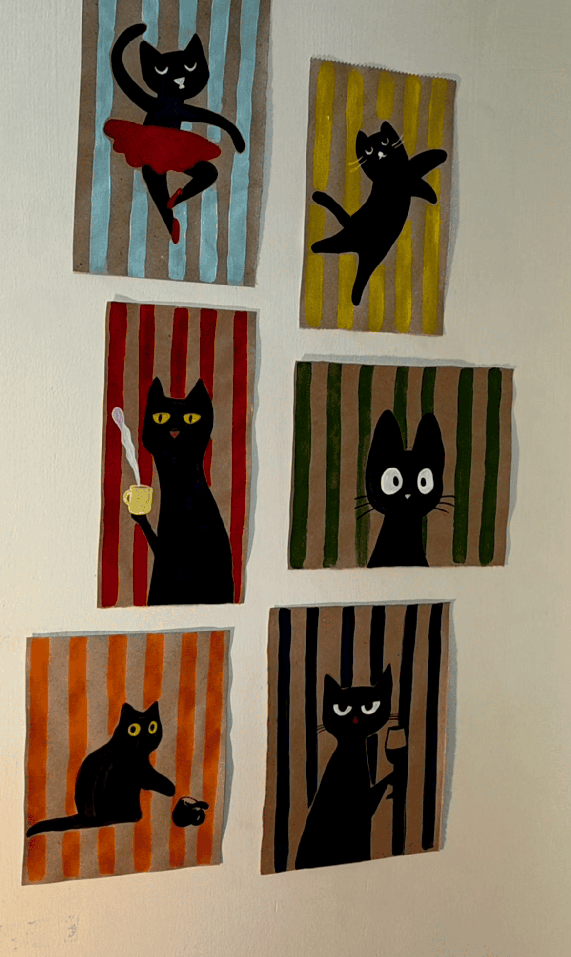 Six small paintings of black cats on striped backgrounds, each cat in a different pose, hanging on a white wall.