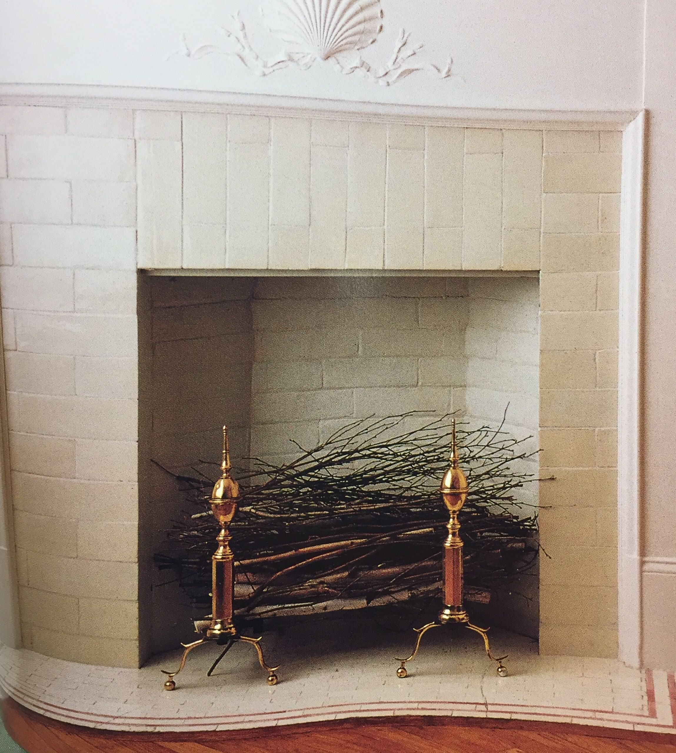 A decorative non-working fireplace filled with bundled branches and two ornate brass andirons.