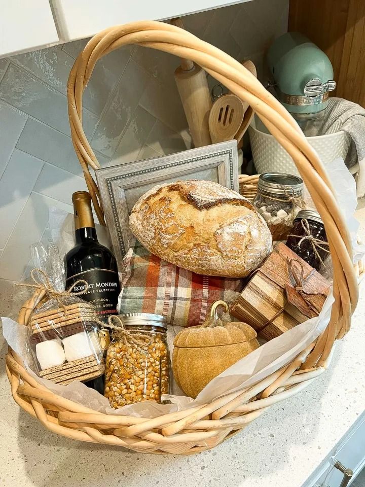 Wicker gift basket filled with a loaf of bread, wine, s'mores kit, popcorn kernels, and a decorative pumpkin.