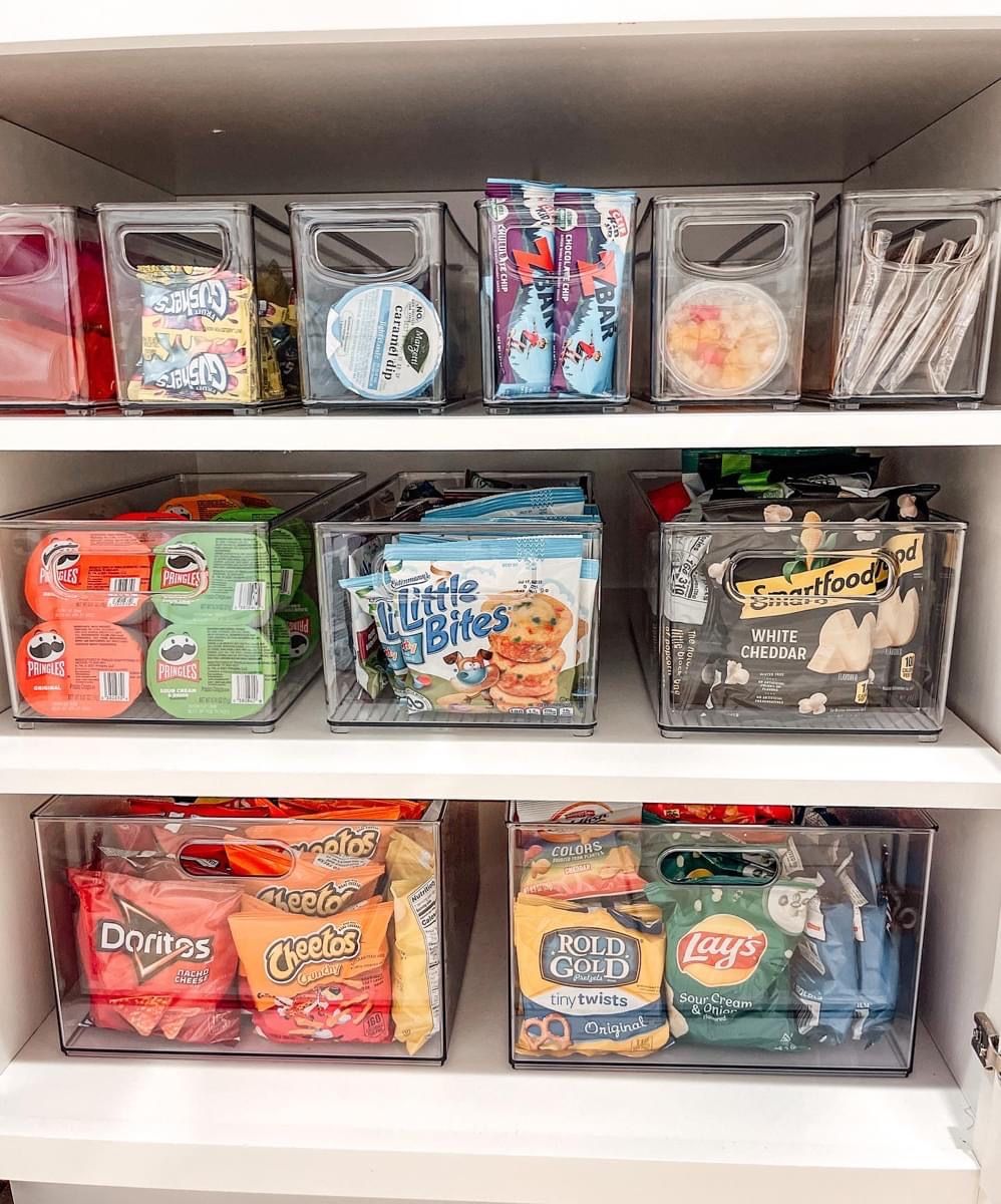White pantry shelves filled with clear bins holding assorted individually packaged snacks and small containers.