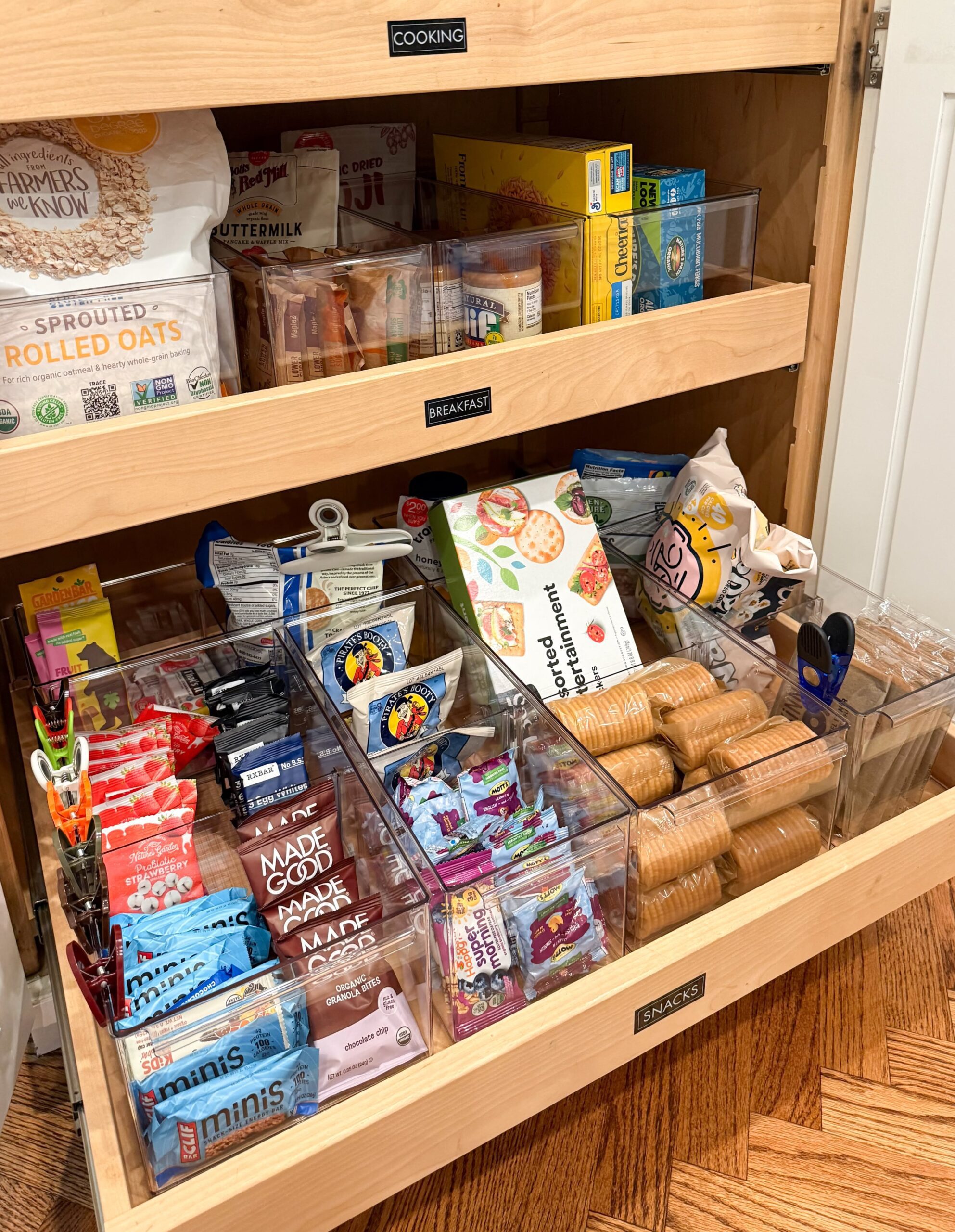 A pantry drawer organized with clear acrylic bins, each holding different types of individually wrapped snacks like bars…