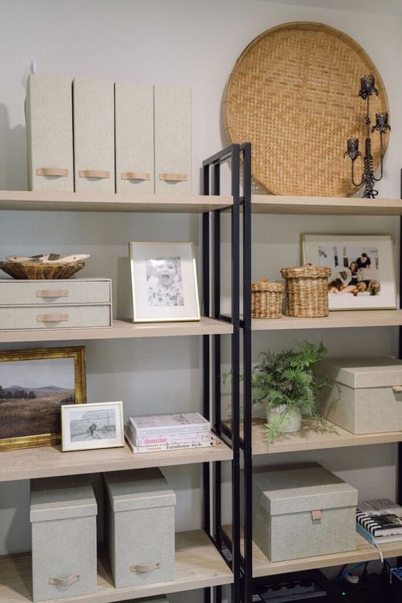 An open bookshelf unit with light wood shelves and black metal frames holds organized bins, magazine files, woven baskets…
