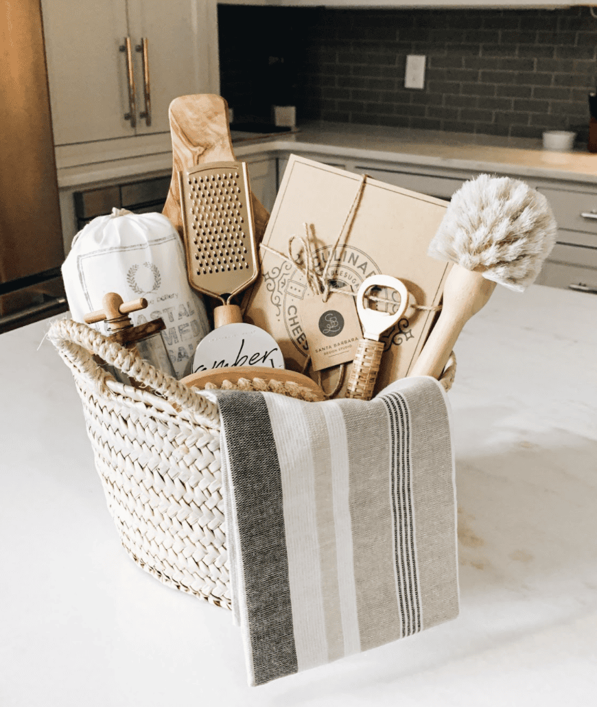 Close-up of a woven basket filled with various kitchen tools, including a gold grater, wooden cheese board, and bottle…