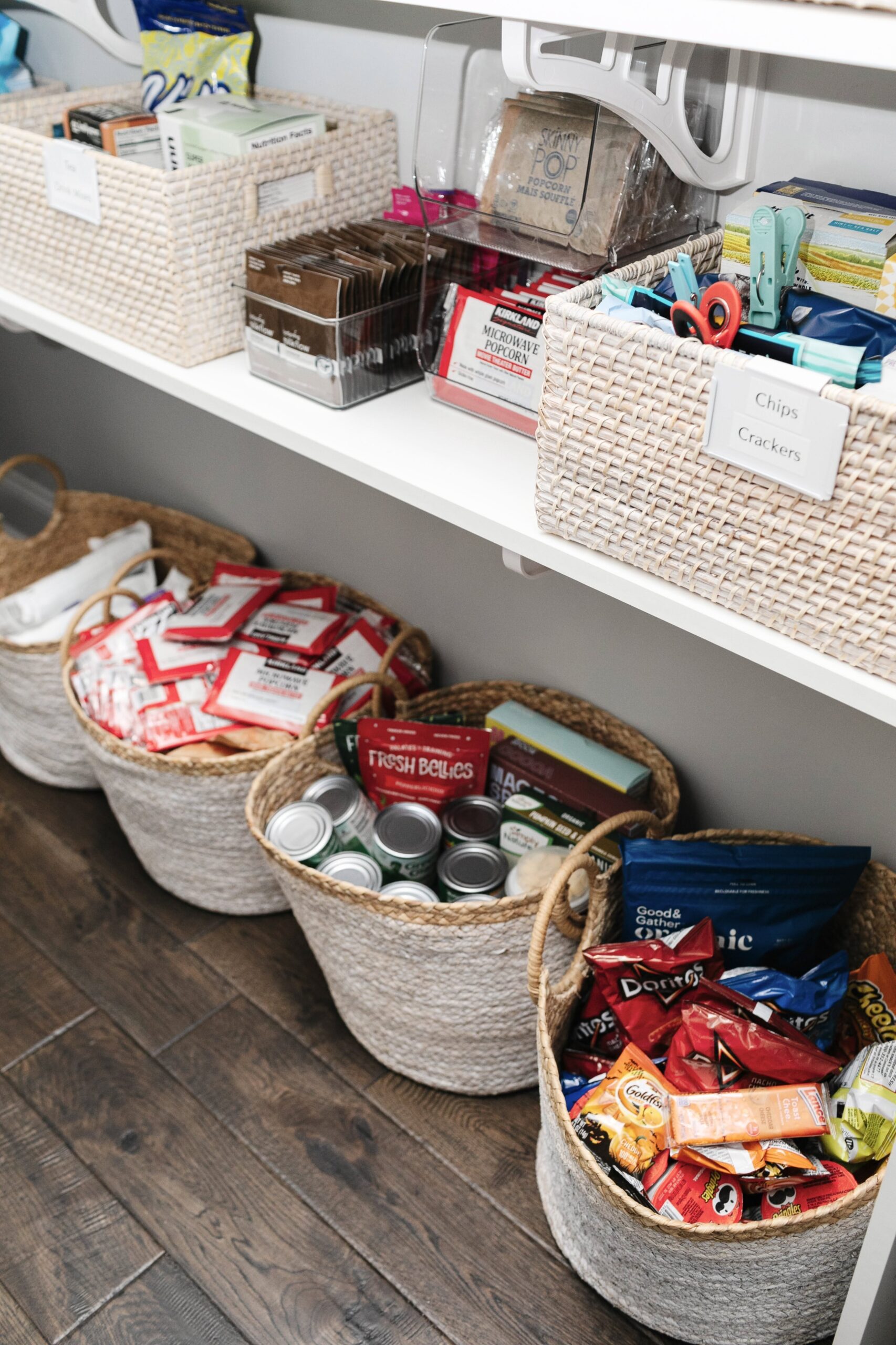 A pantry shelf with woven baskets organized with various snack foods, including chips, popcorn, and other packaged items.