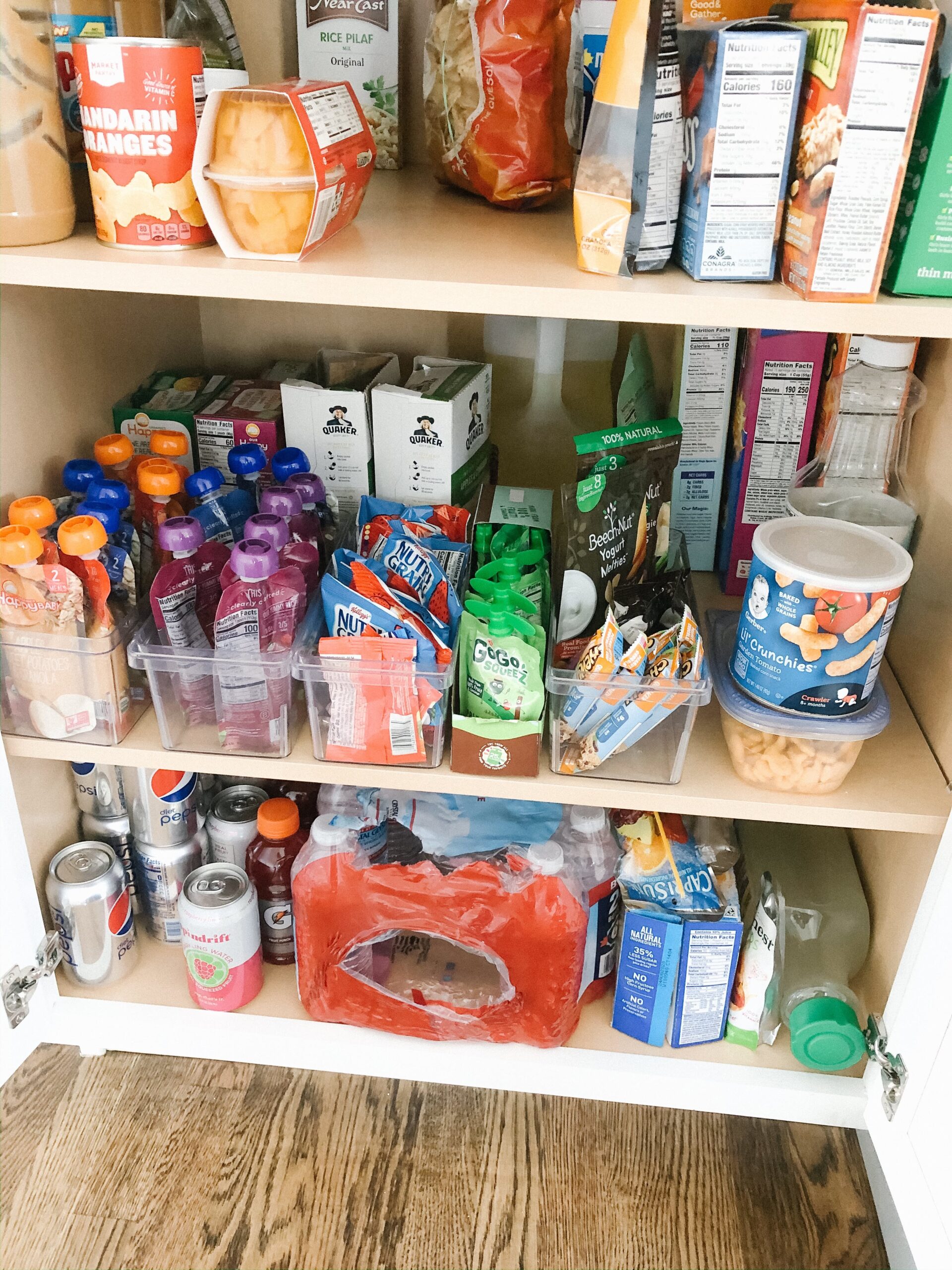 A pantry shelf with various snacks in clear plastic bins, including baby food pouches, fruit purees, and cracker snacks.