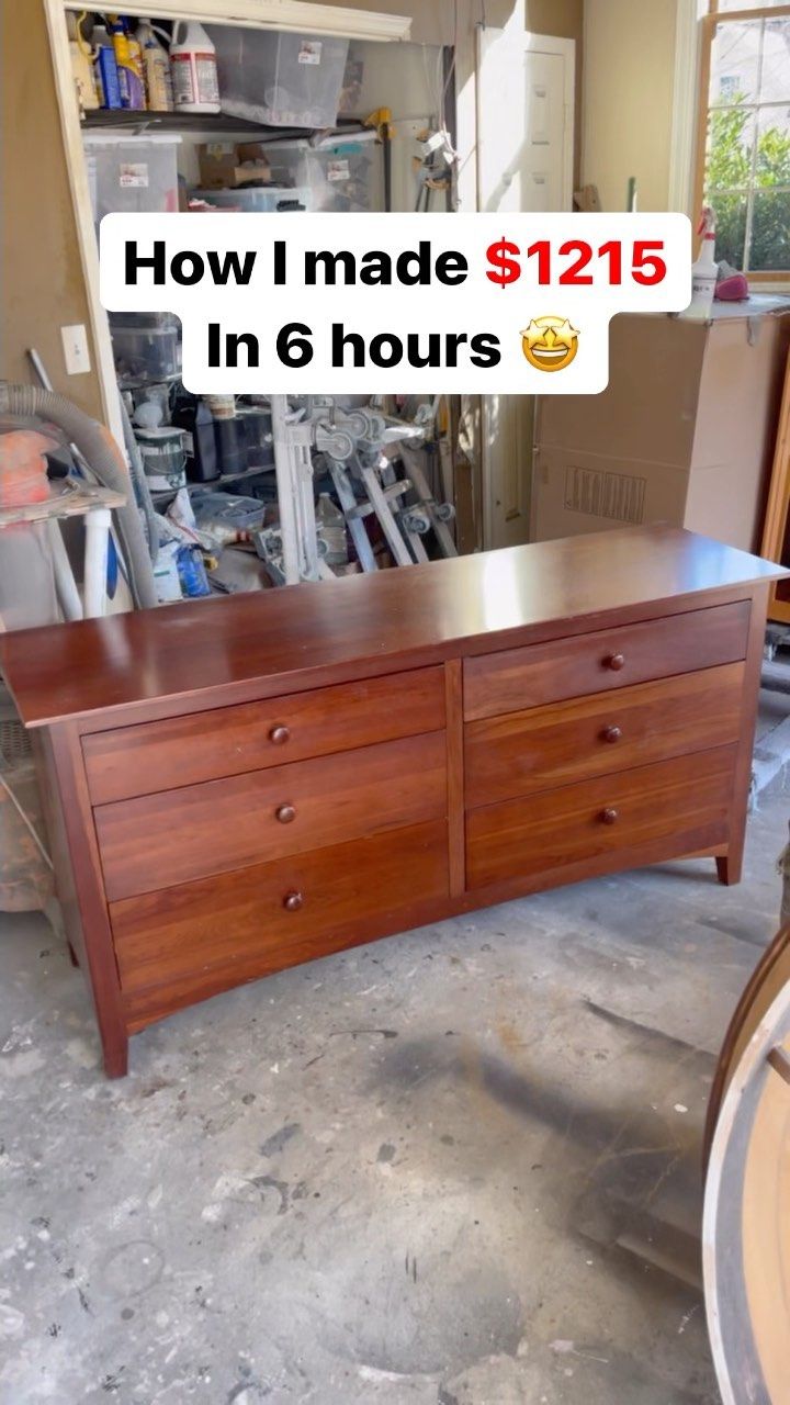 A refurbished wooden dresser with six drawers sits in a workshop with tools and supplies visible in the background.