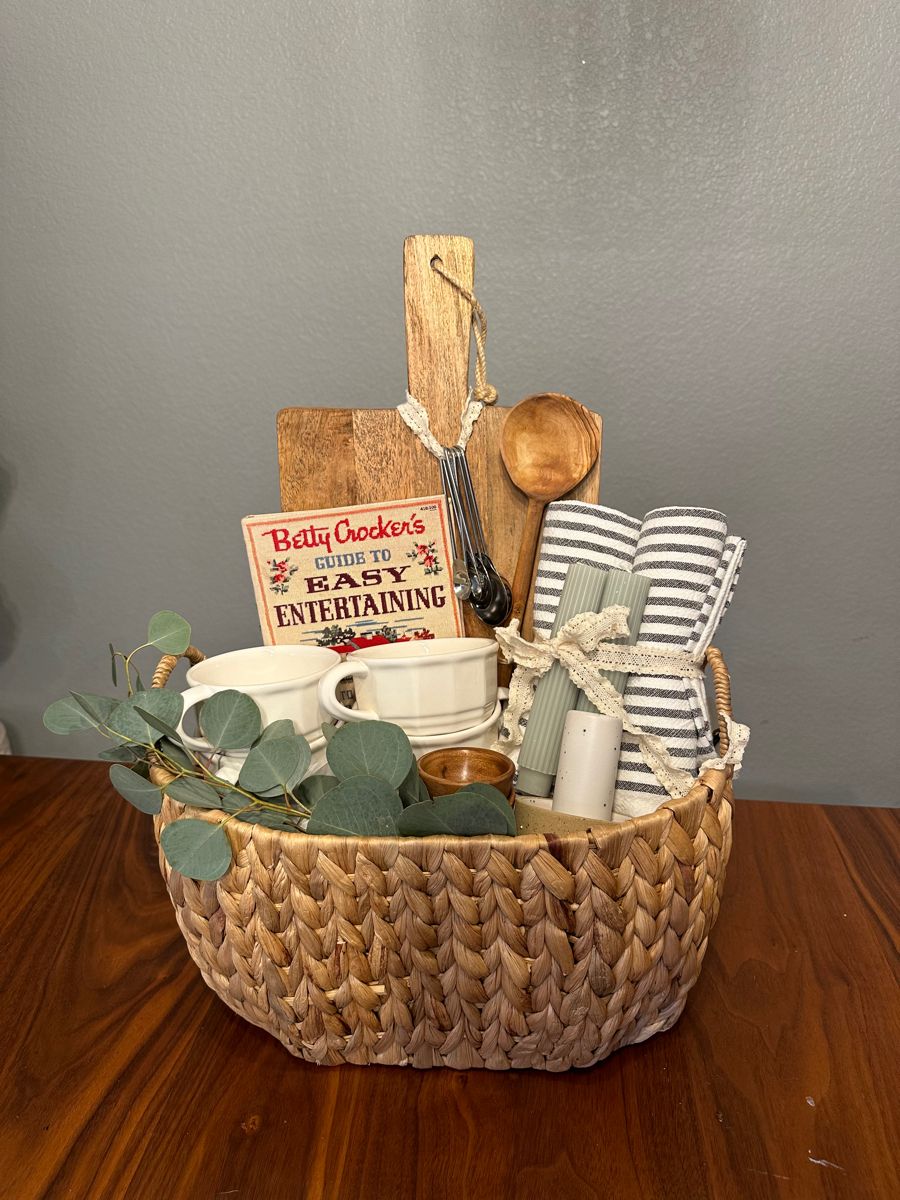 Woven basket filled with small white bowls and cups, kitchen utensils, striped napkins, a cookbook, and green eucalyptus…