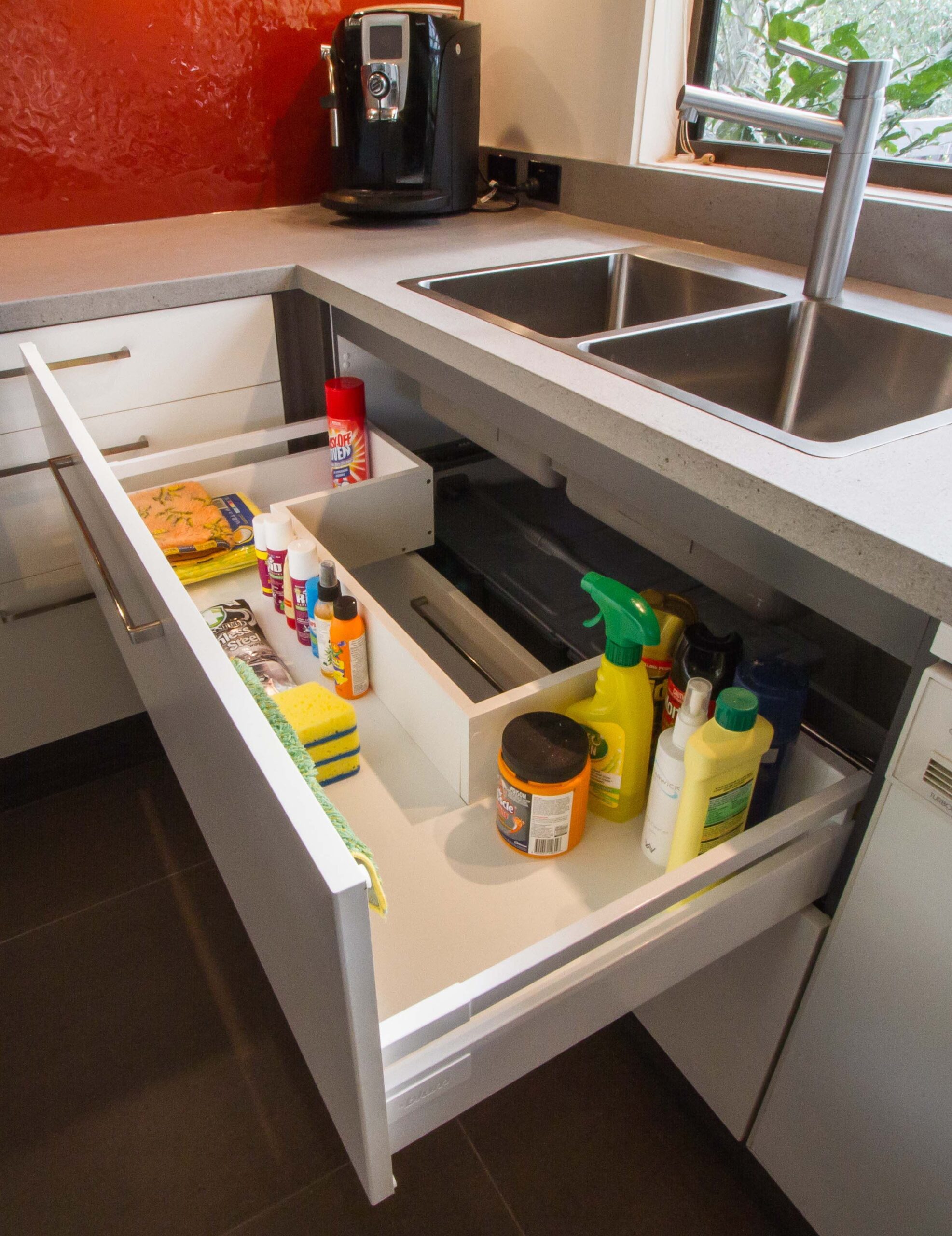 Open kitchen drawer under a sink, custom-built with cutouts for pipes, holding various cleaning supplies.
