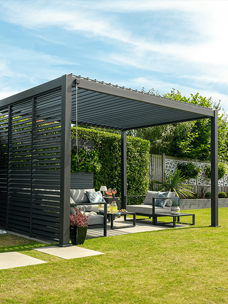 Modern dark gray pergola with adjustable louvered roof and side privacy screen, covering an outdoor lounge set in a garden.