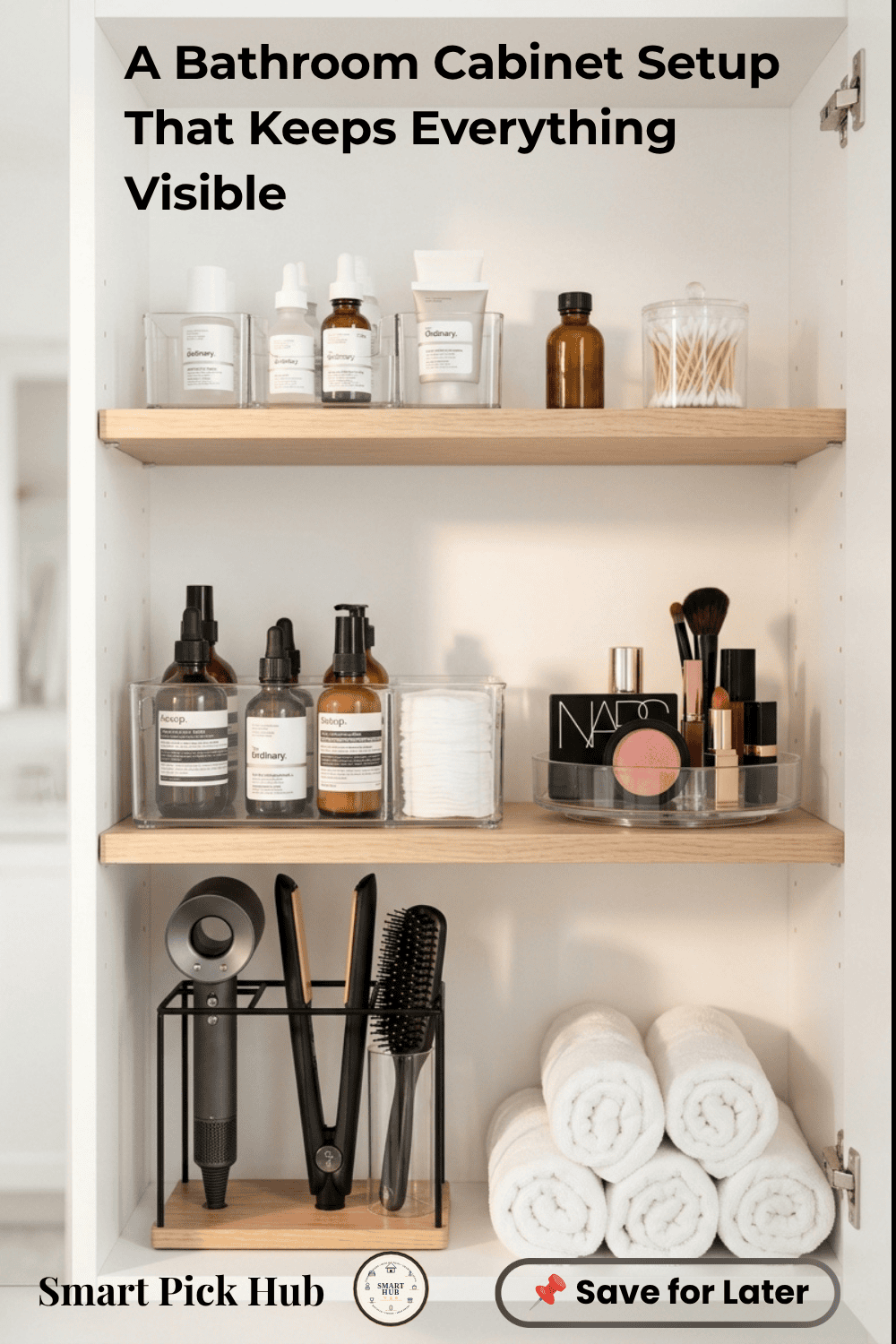 Open bathroom cabinet with wooden shelves holding clear containers of skincare, makeup, hair tools, and rolled towels.
