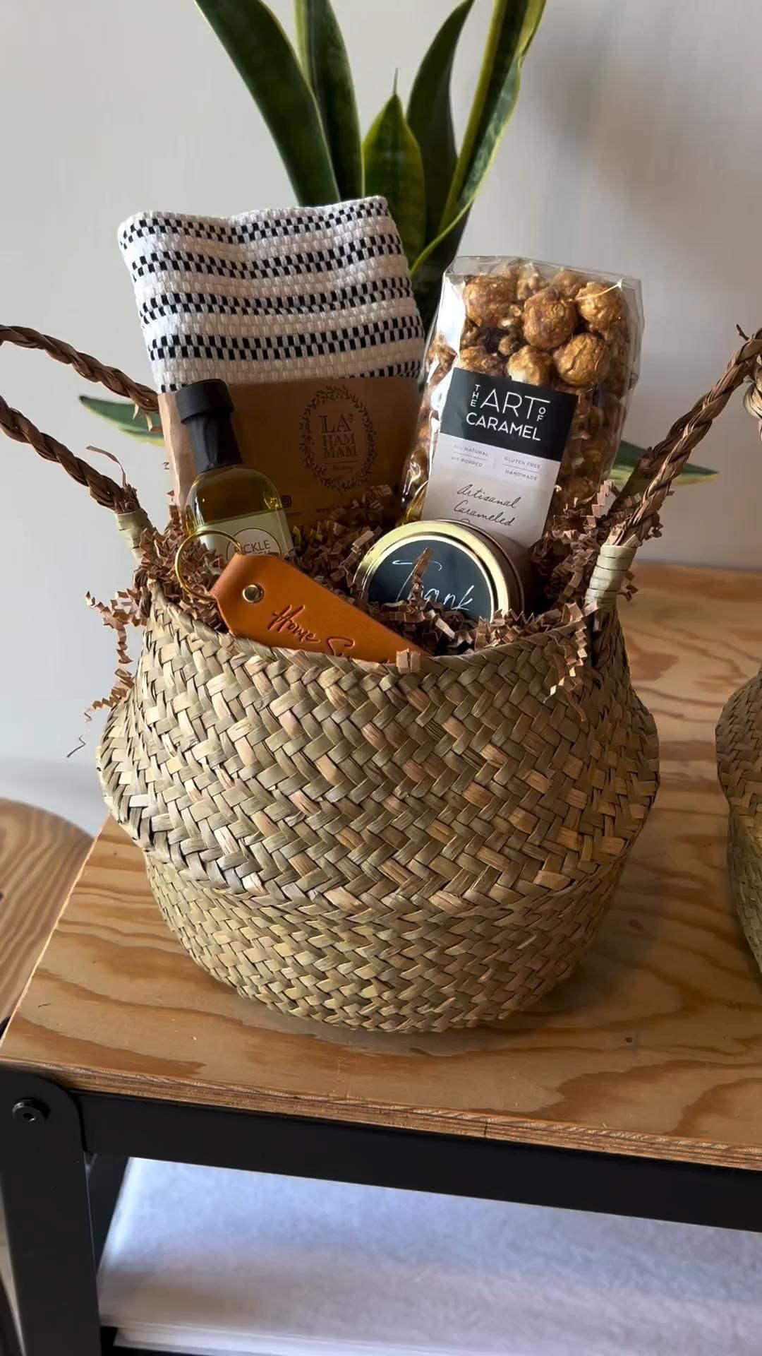Woven basket filled with gourmet popcorn, olive oil, a small striped towel, a candle, and a leather tag with 'Home' embossed…