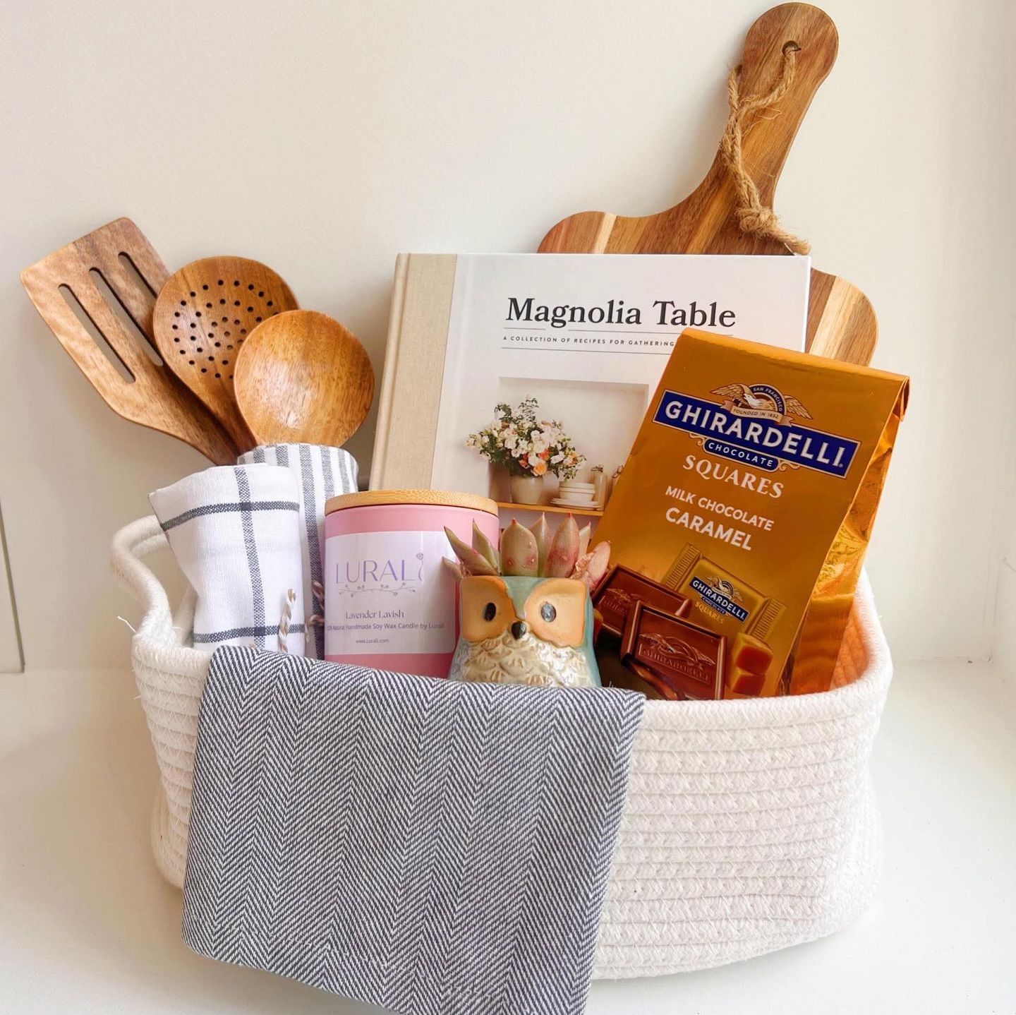 A white rope basket filled with wooden kitchen utensils, dish towels, a cookbook, a candle, a small succulent, and…