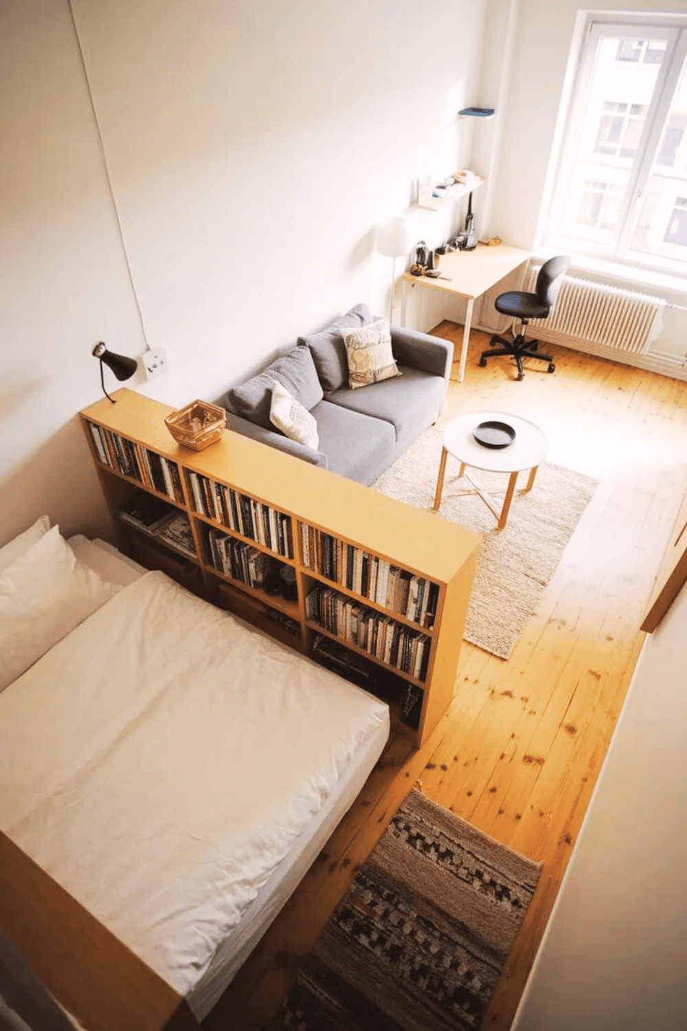 Overhead view of a studio apartment divided by a wooden bookcase, separating a bed from a gray sofa, coffee table, and desk.