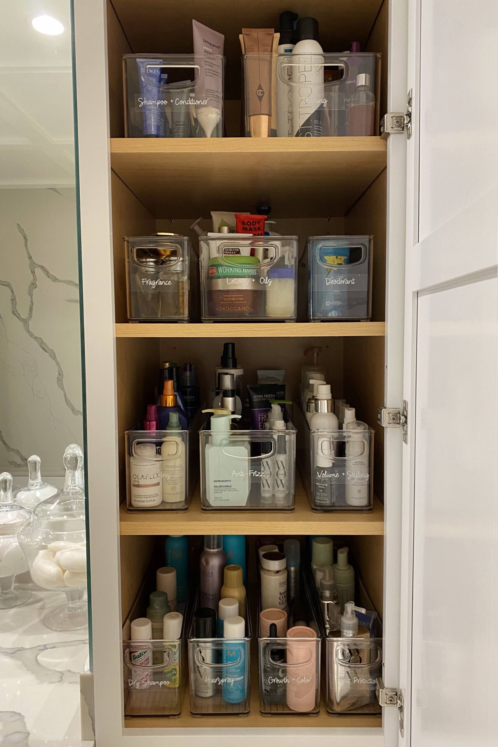 Inside a wood-shelved bathroom cabinet, clear bins organize various beauty and personal care products.