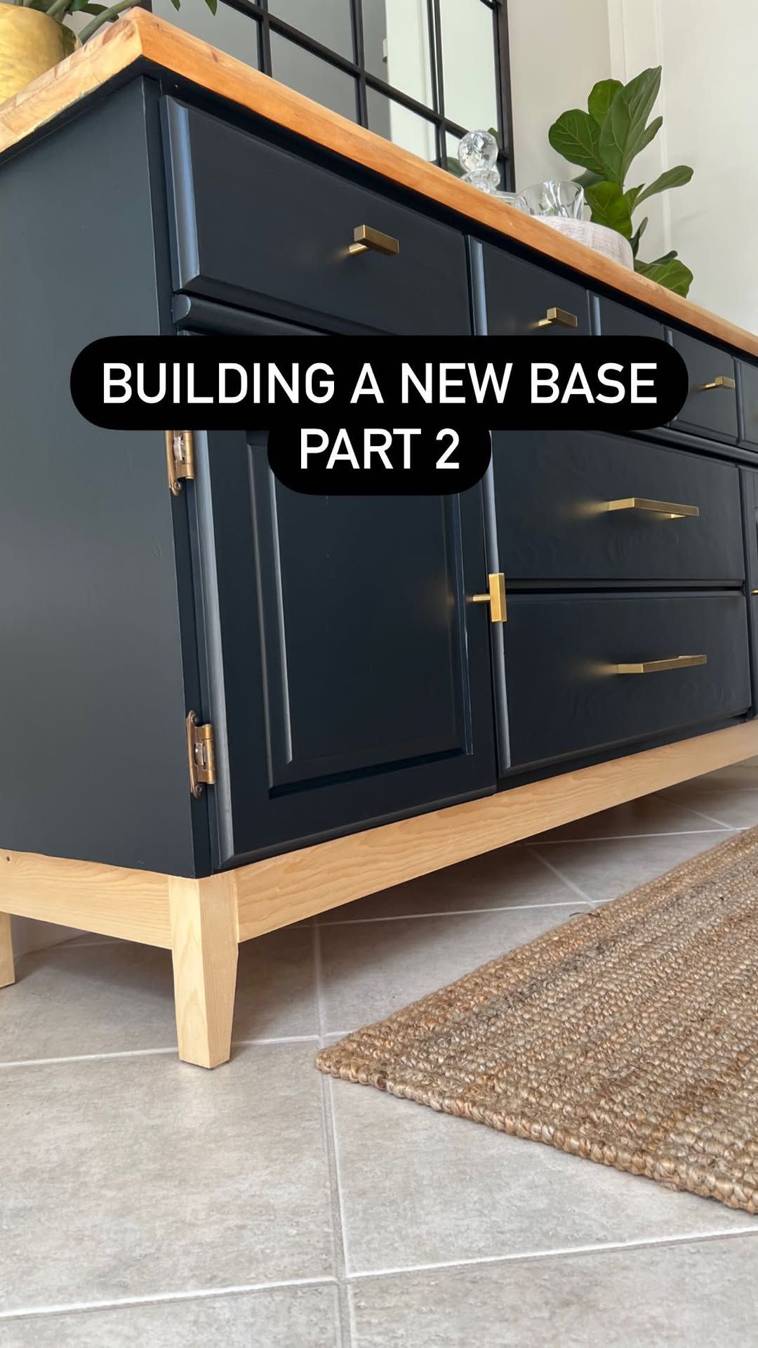 Dark blue dresser with gold hardware and a light wood custom base, on a tiled floor next to a woven rug.