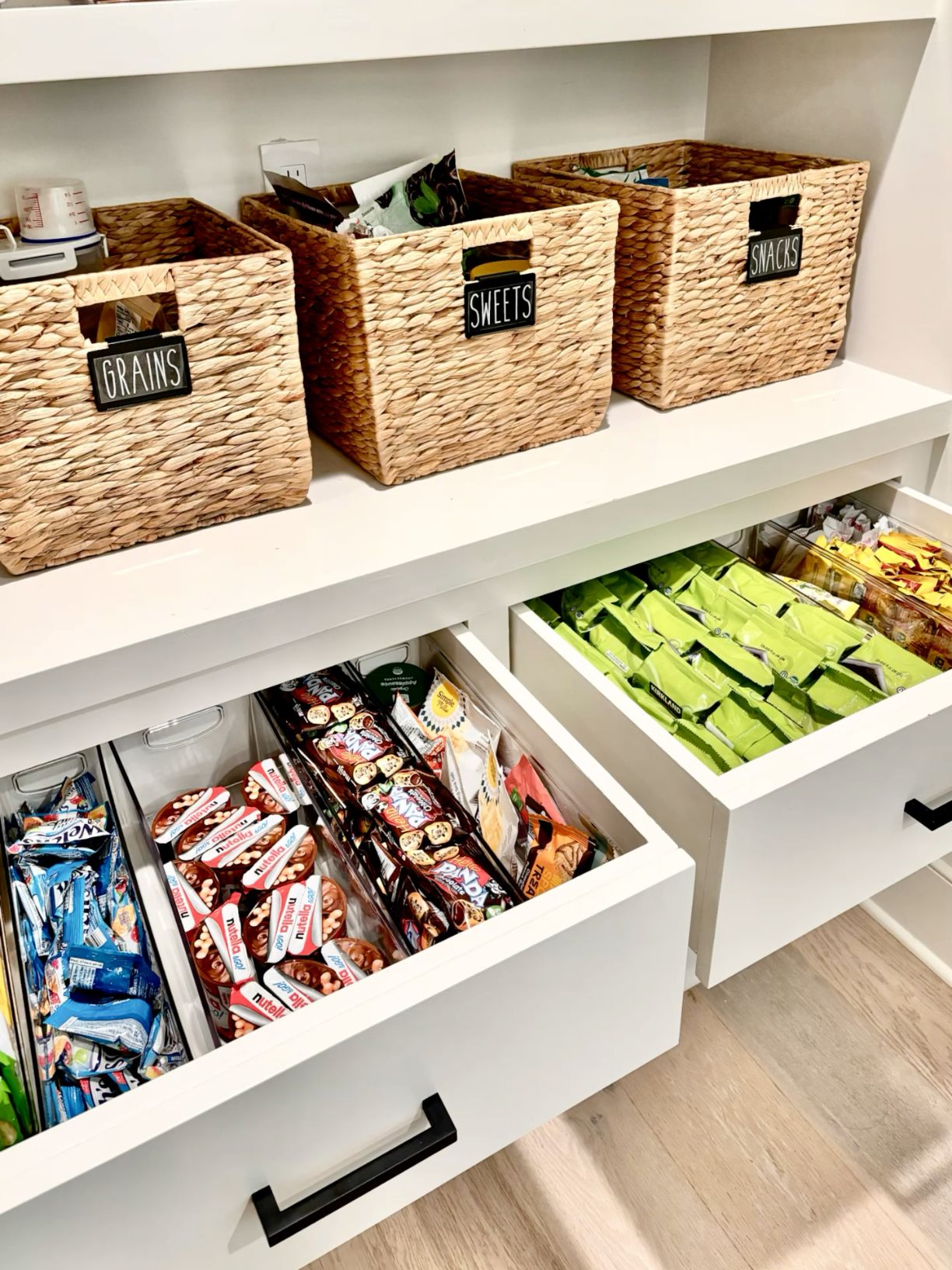 Pantry with organized shelves and open drawers. Top shelves hold labeled woven baskets for 'Grains', 'Sweets', and 'Snacks'.