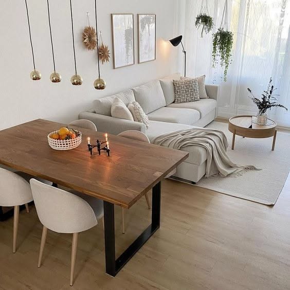 A small apartment living room with a cream sectional sofa, a round coffee table, and an adjacent wooden dining table with…