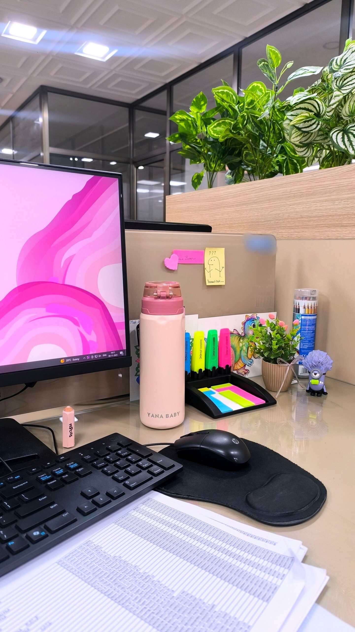 A desk with a computer monitor, keyboard, mouse, water bottle, highlighters, and a small potted plant on the right side.