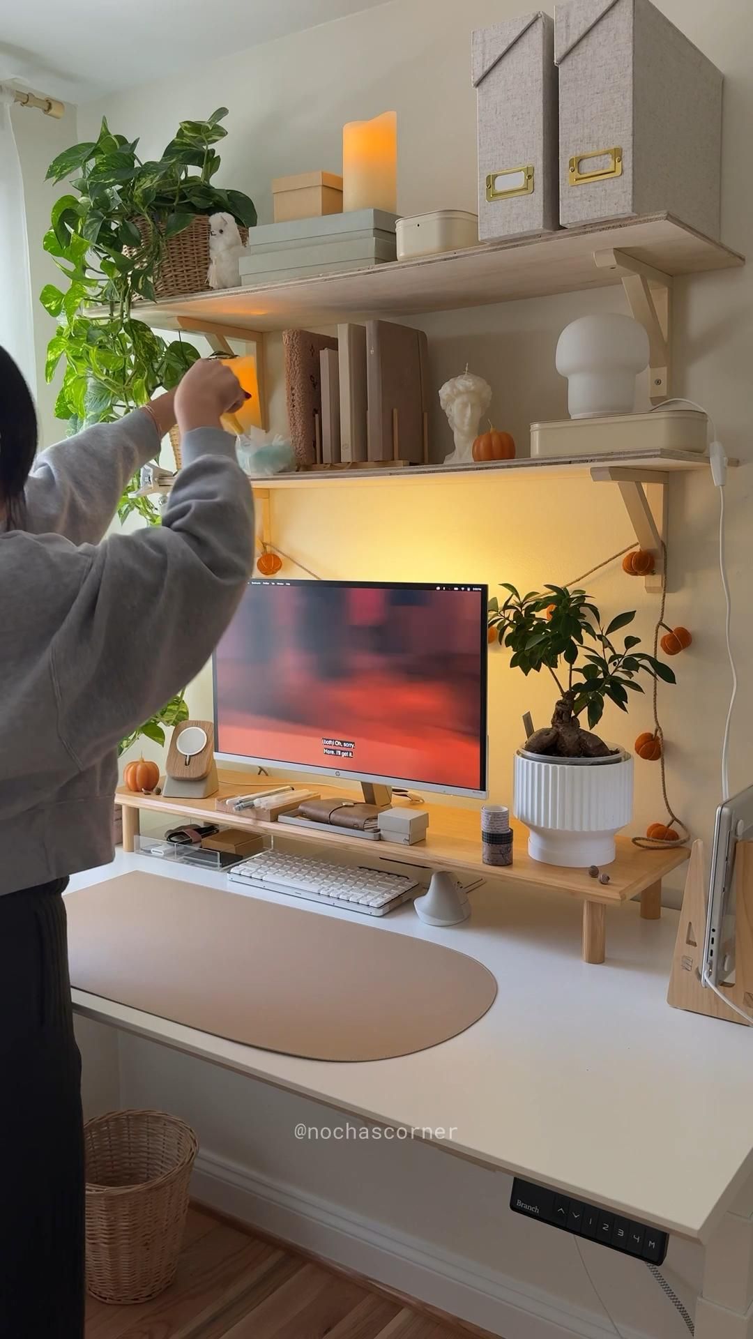 A person stands at a light-toned wood desk, arranging items on a shelf above a computer monitor, illuminated by a warm light.