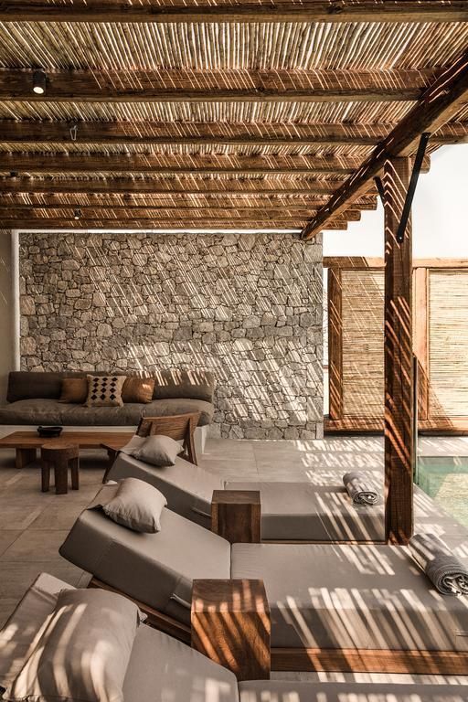 An outdoor living area featuring a stone wall, wooden pergola with woven reed roofing, lounge chairs, and a sectional sofa.