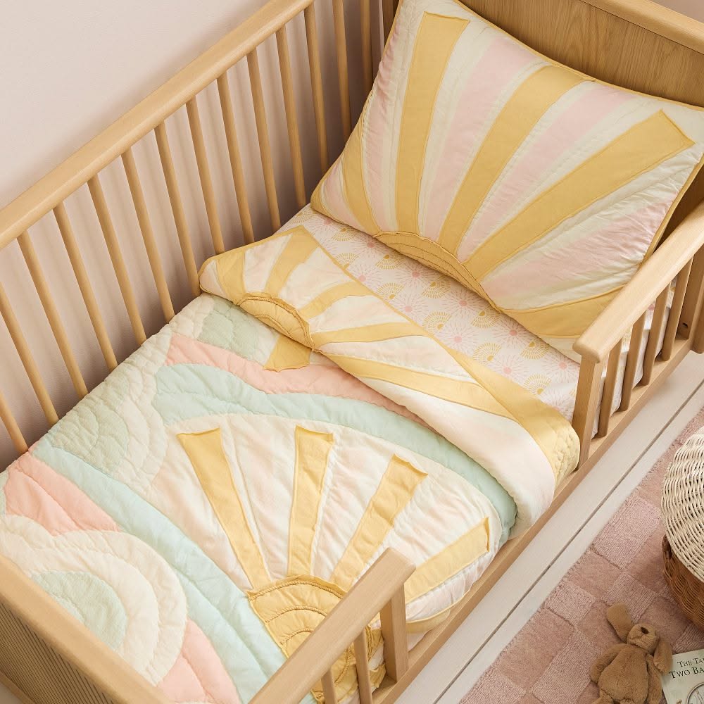 Crib with a light wood frame holding a quilt and pillow with a yellow, pink, and mint green sunburst pattern