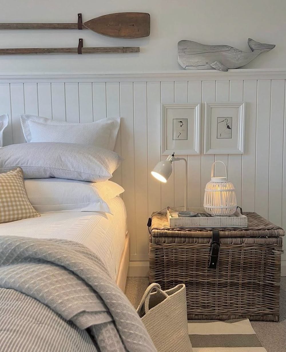 A bedroom with white paneling and a bed with light-colored bedding. A woven trunk serves as a bedside table.