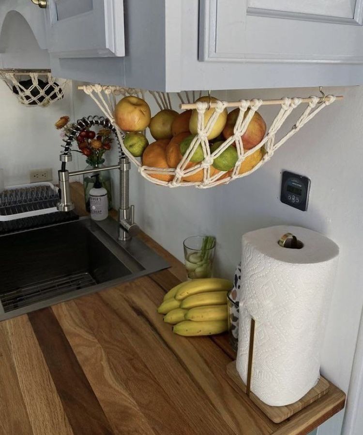 Macrame hammocks filled with various fruits hanging beneath light gray kitchen cabinets over a wooden counter.