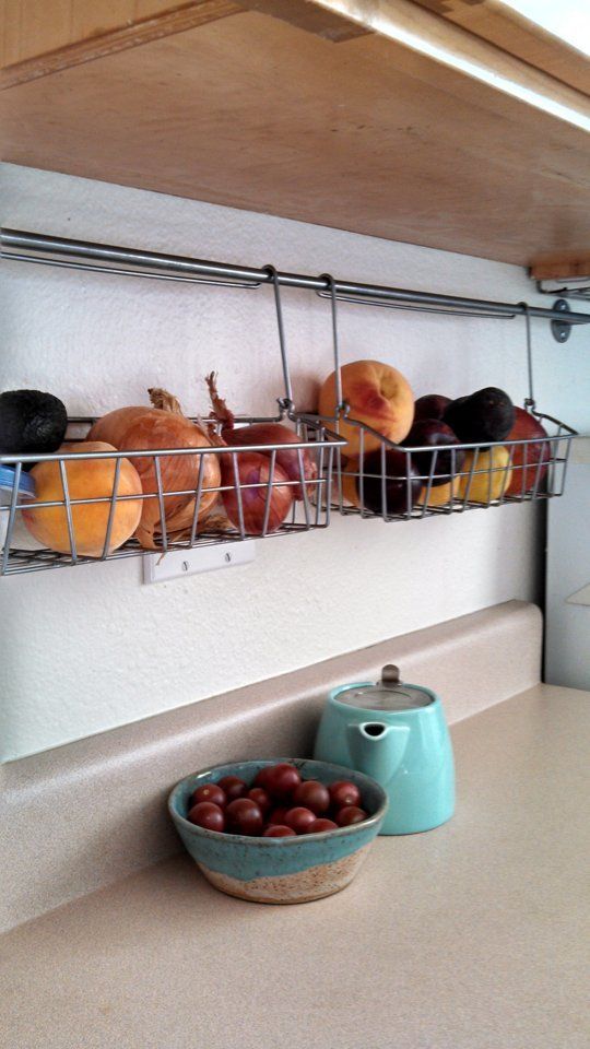 Wire baskets hanging from a rod under a wooden cabinet, filled with onions, peaches, and plums.