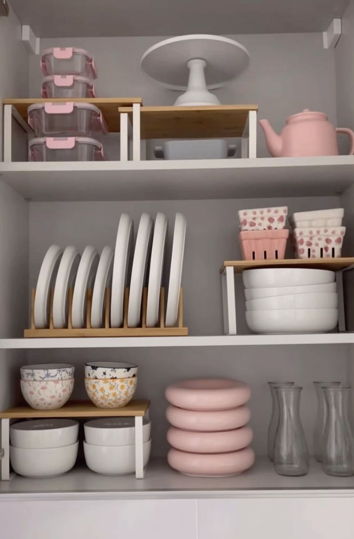 Open kitchen cabinet shelves organized with risers, holding plates, bowls, a teapot, and food storage containers.