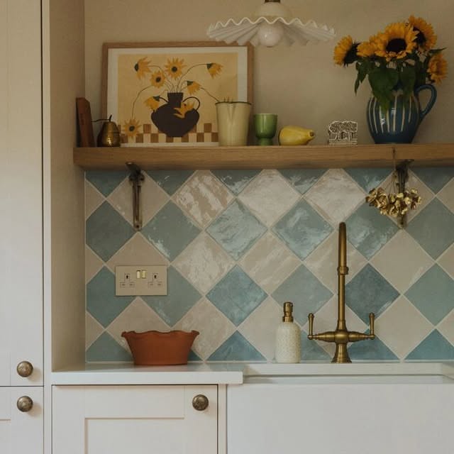 Kitchen backsplash with light blue and white square tiles laid diagonally above a white sink and brass faucet.