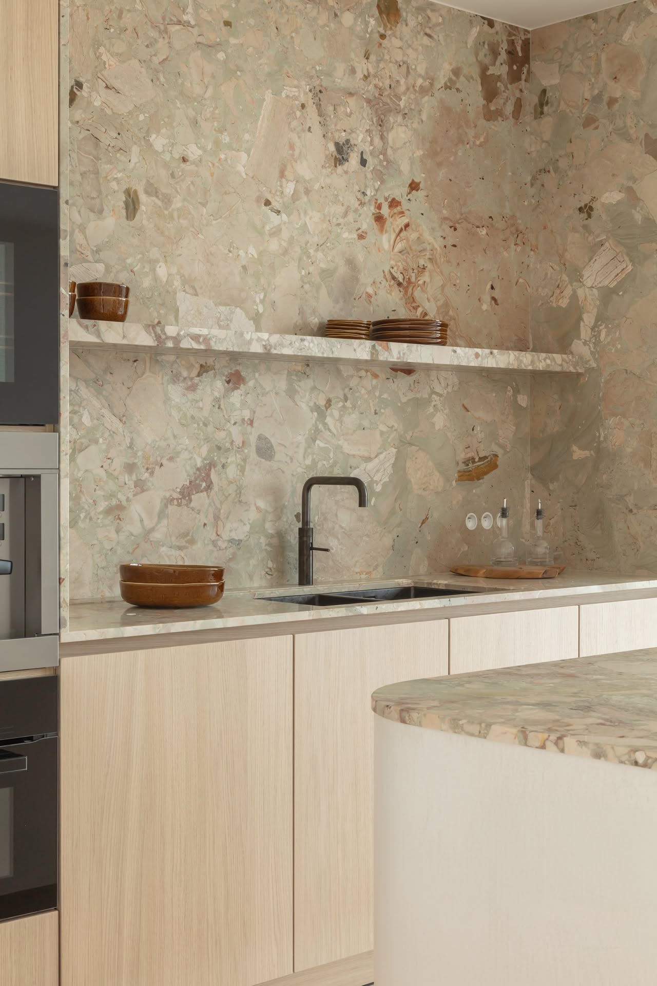 Kitchen with light wood cabinets, black faucet, and a variegated stone backsplash that extends up to the ceiling with…